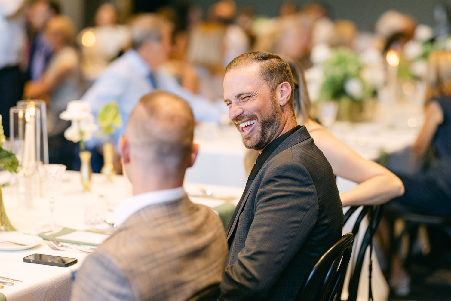 A person laughing heartily while sitting at a decorated banquet table with other guests in soft focus in the background.