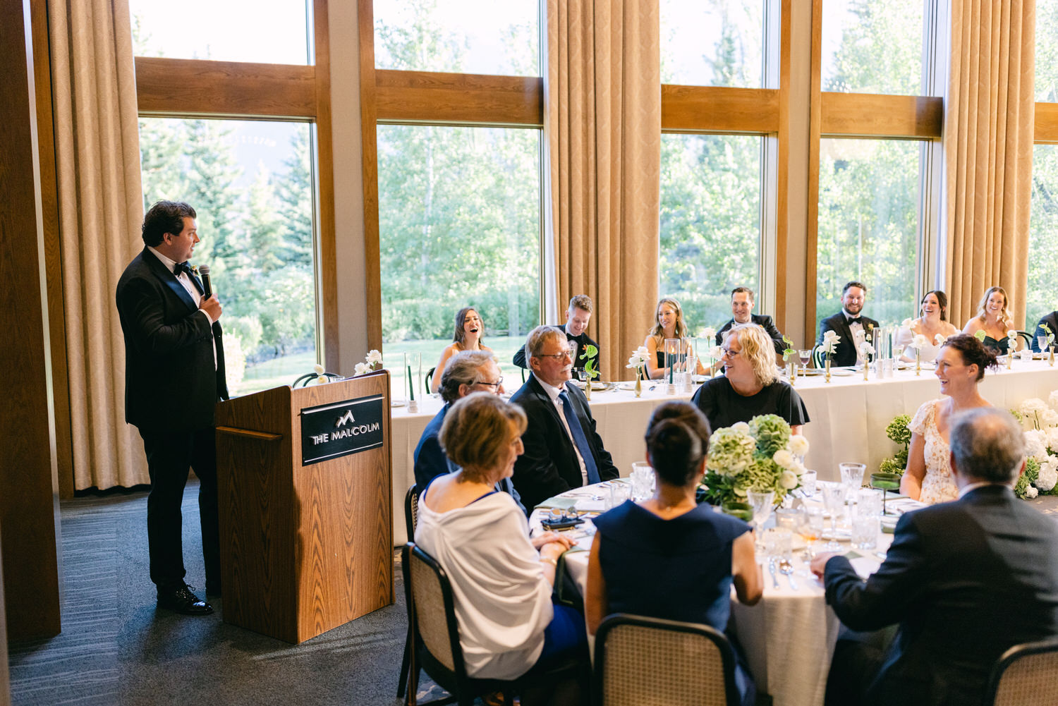 A man in a tuxedo giving a speech at a wedding reception while guests listen attentively