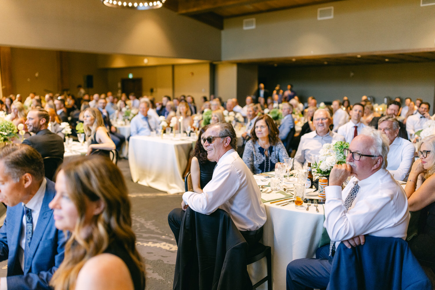 Audience seated at decorated tables during a formal indoor event, with some guests looking towards a focal point off-camera.