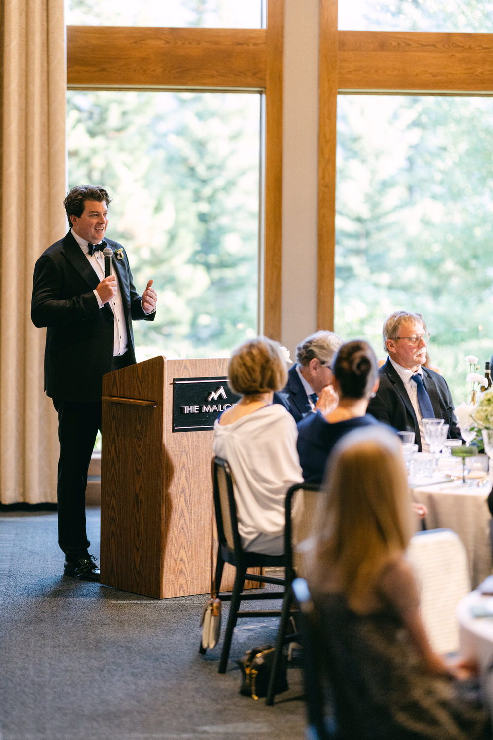 A man in a tuxedo speaking at a podium with guests seated at the dinner table, in an event hall with a view of trees outside the window.