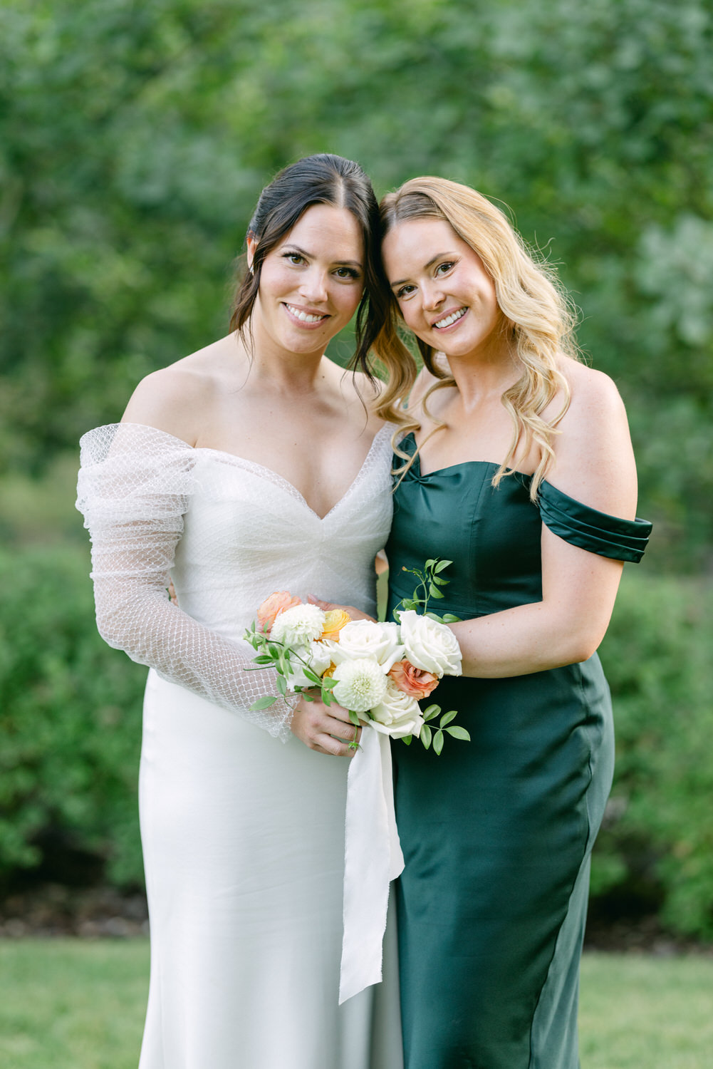 Two women posing for a photo, one in a white wedding dress with a bouquet and the other in a dark green dress, with a natural backdrop.