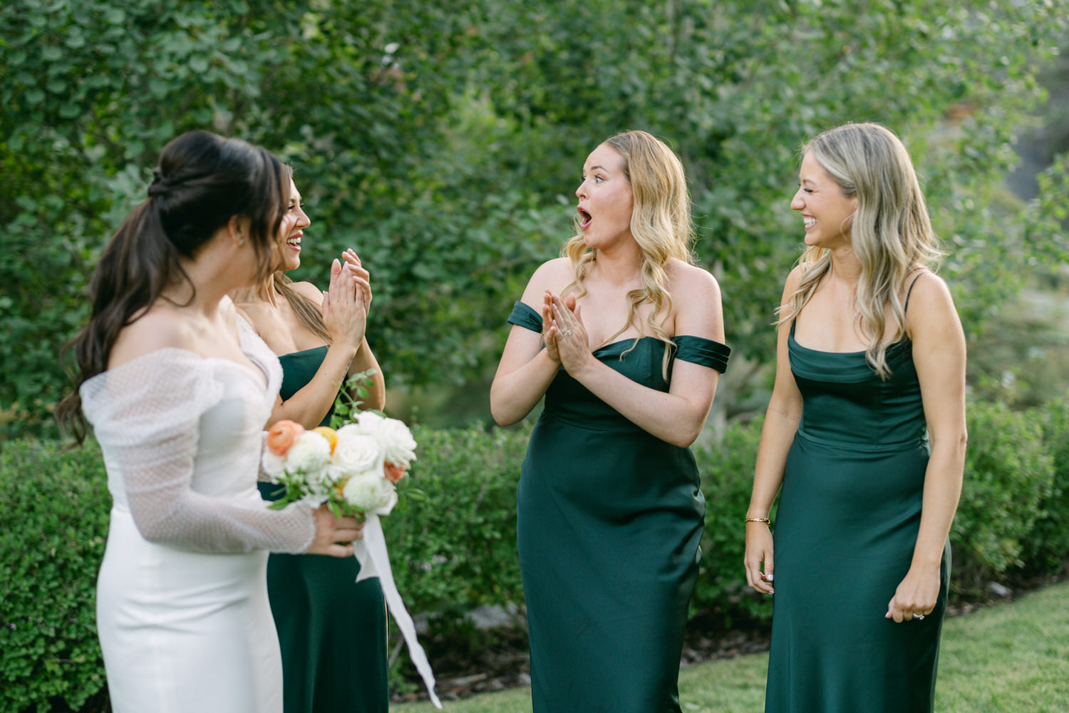 Three women in dark green dresses express excitement and joy with a bride wearing a white off-the-shoulder dress holding a bouquet of flowers, in an outdoor wedding setting.