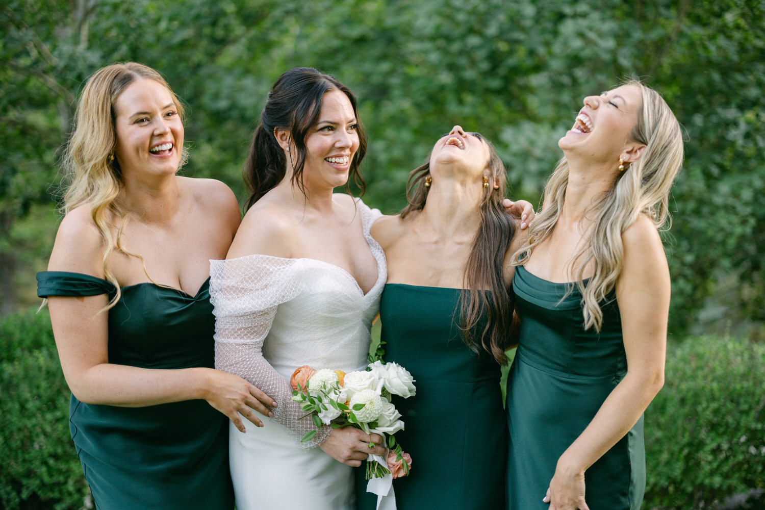 A bride in a white dress laughing with three bridesmaids in dark green dresses, all enjoying a cheerful moment together outdoors.