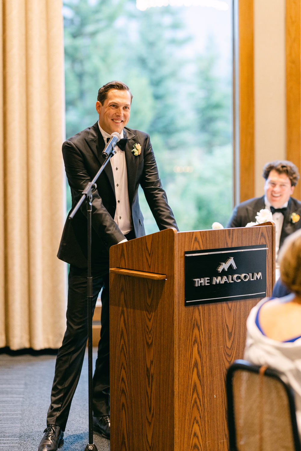 A smiling man in a tuxedo speaking at a microphone on a podium with 'The Malcolm' sign, with a guest laughing in the background