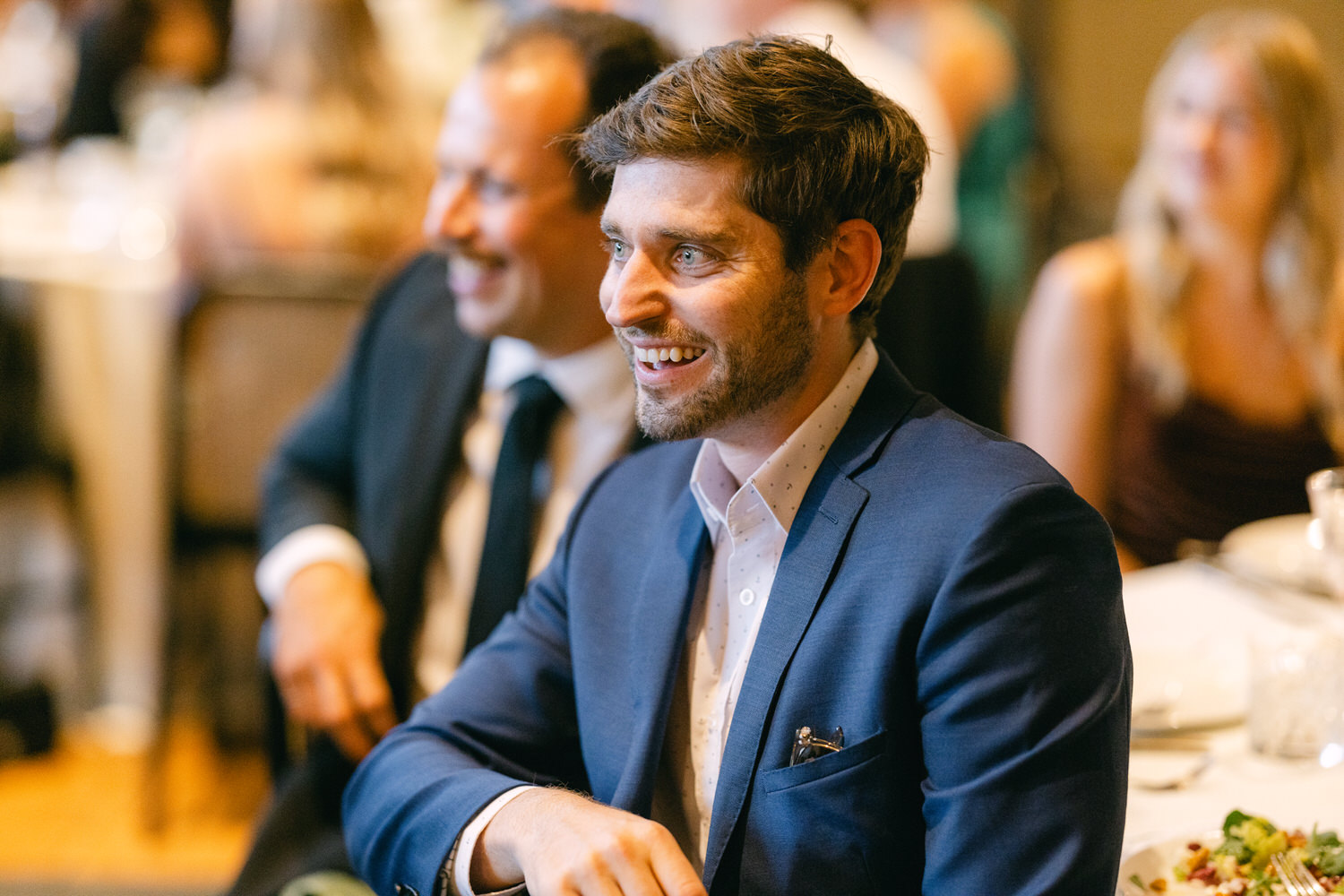 A man in a blue suit with a beaming smile at a social gathering, surrounded by other guests enjoying the occasion.