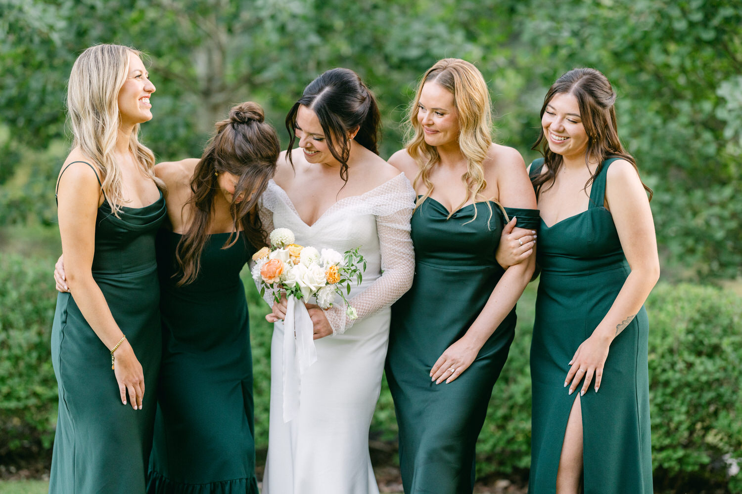 A bride in white standing with her bridesmaids dressed in green, laughing and enjoying a special occasion outdoors.