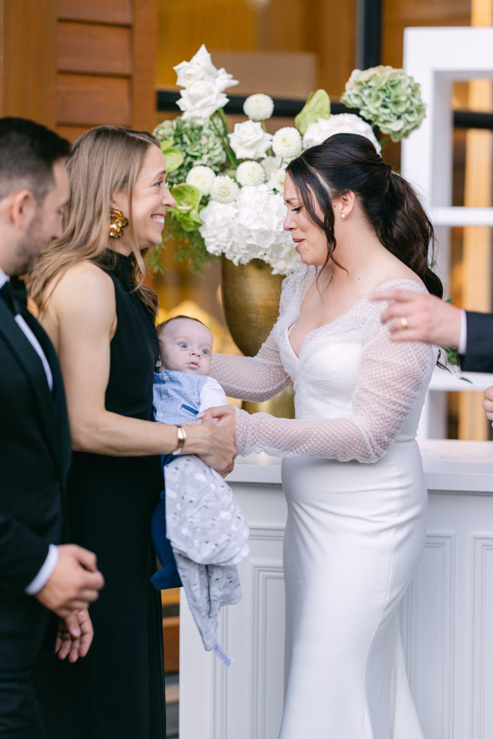 A bride holding a baby interacts warmly with a smiling woman while others look on, against a backdrop of white floral arrangements.