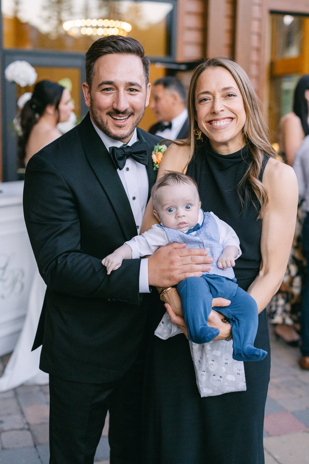 A man in a tuxedo and a woman in a black dress smiling for the camera, holding a baby in a blue outfit.