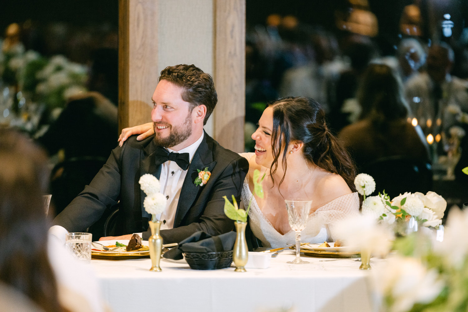 A man and a woman smiling and enjoying themselves at a wedding reception table, surrounded by elegant white floral arrangements and dinner settings