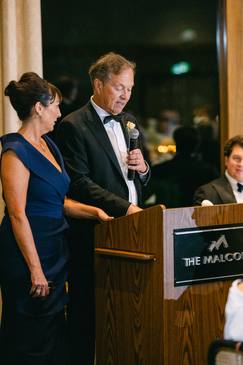 A man in a tuxedo speaks at a lectern beside a woman in a blue dress at an elegant indoor event.