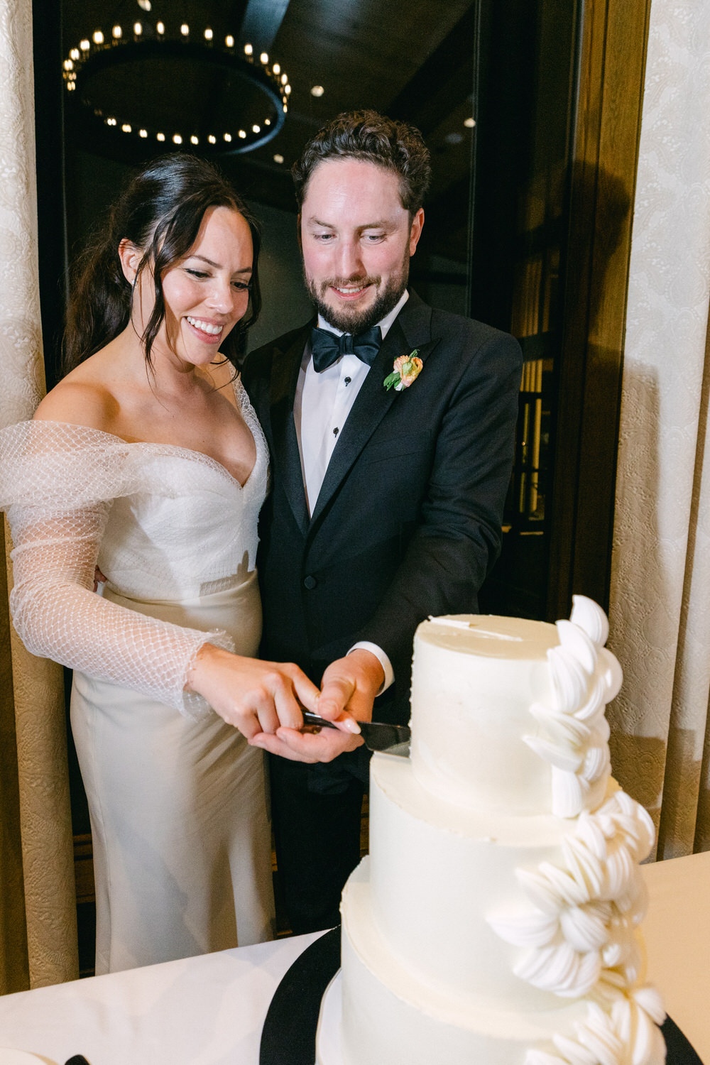 A happy couple in wedding attire cutting a multi-tiered white wedding cake together.