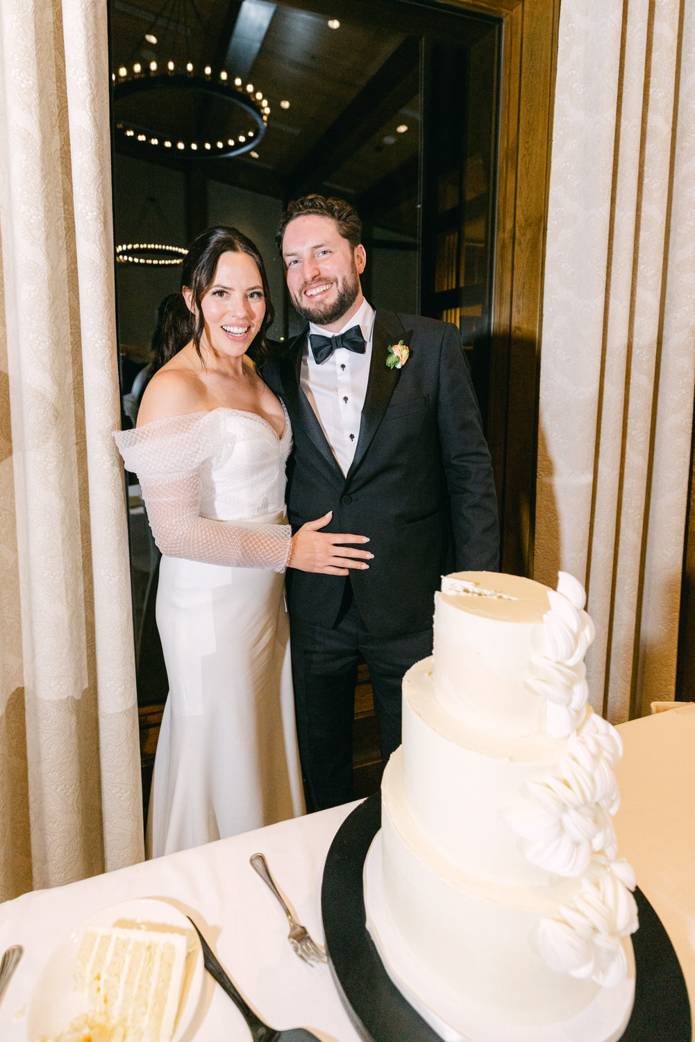 A happy couple in wedding attire smiling beside a three-tiered wedding cake in a decorated venue.