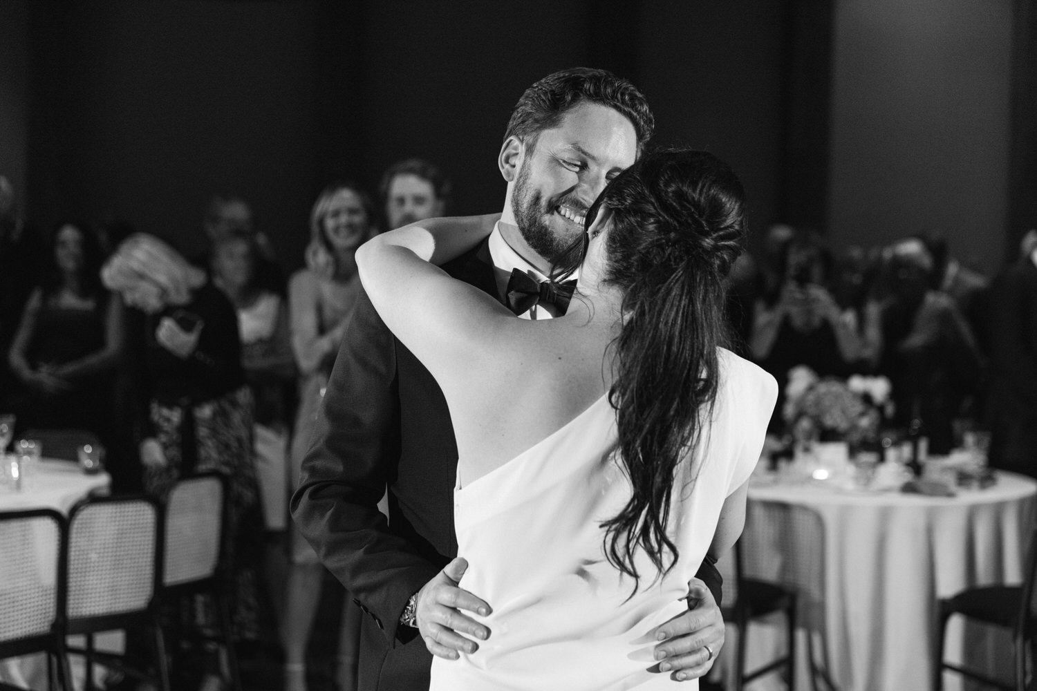 A monochrome image of a bride and groom sharing their first dance, surrounded by guests at their wedding reception.