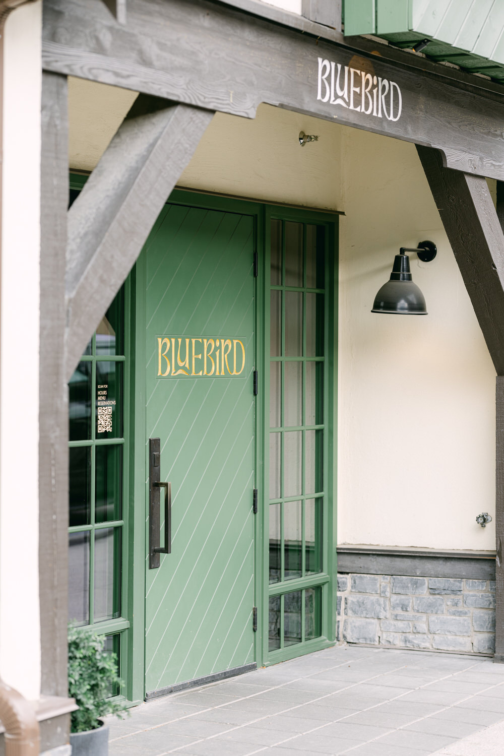 A green door of a building labeled "BLUEBIRD" under a wooden beam with stone accents and an outdoor lamp overhead.