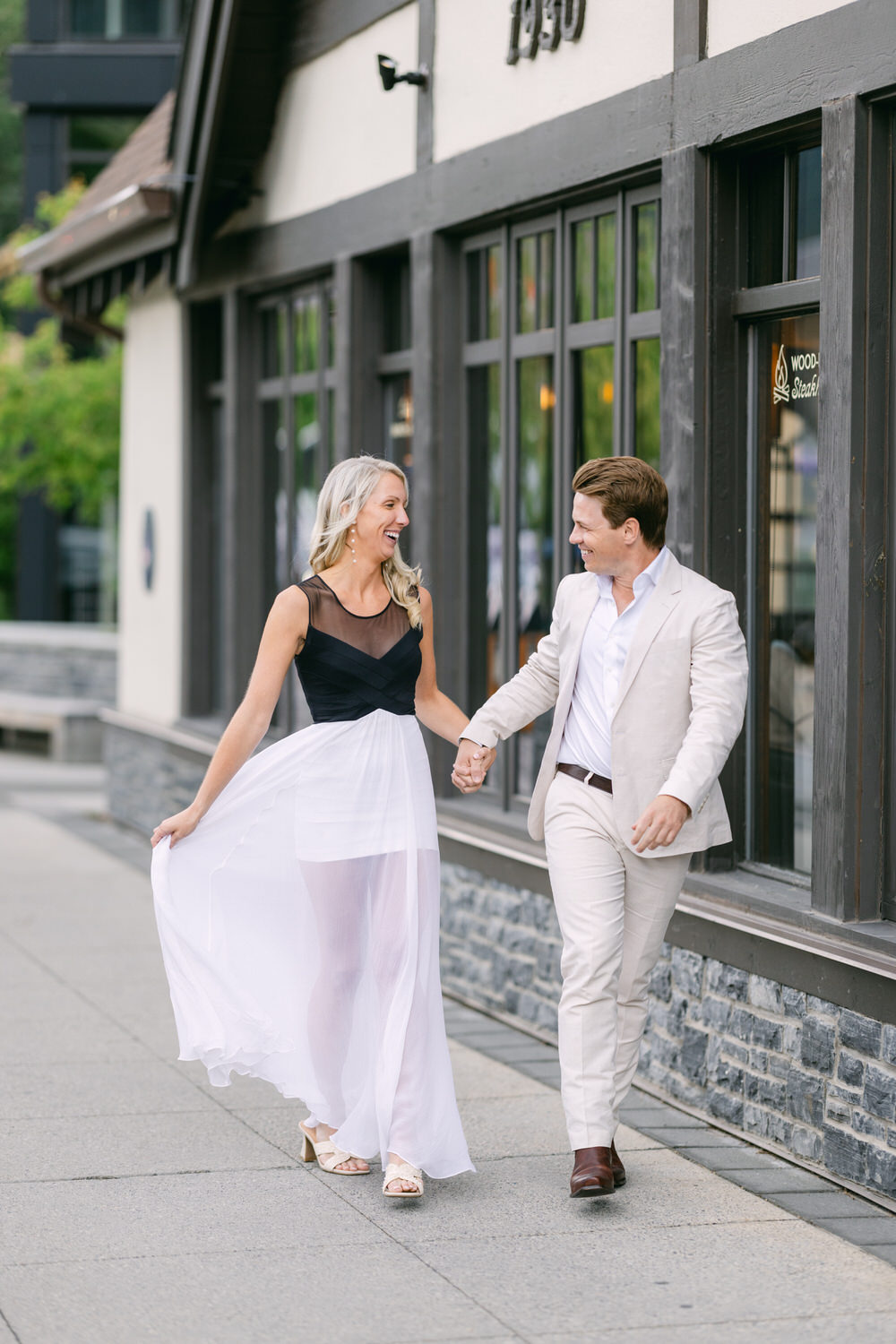 A smiling man and woman holding hands and walking along a city sidewalk, with the woman wearing a white flowing skirt and black top, and the man in a white jacket and beige trousers.