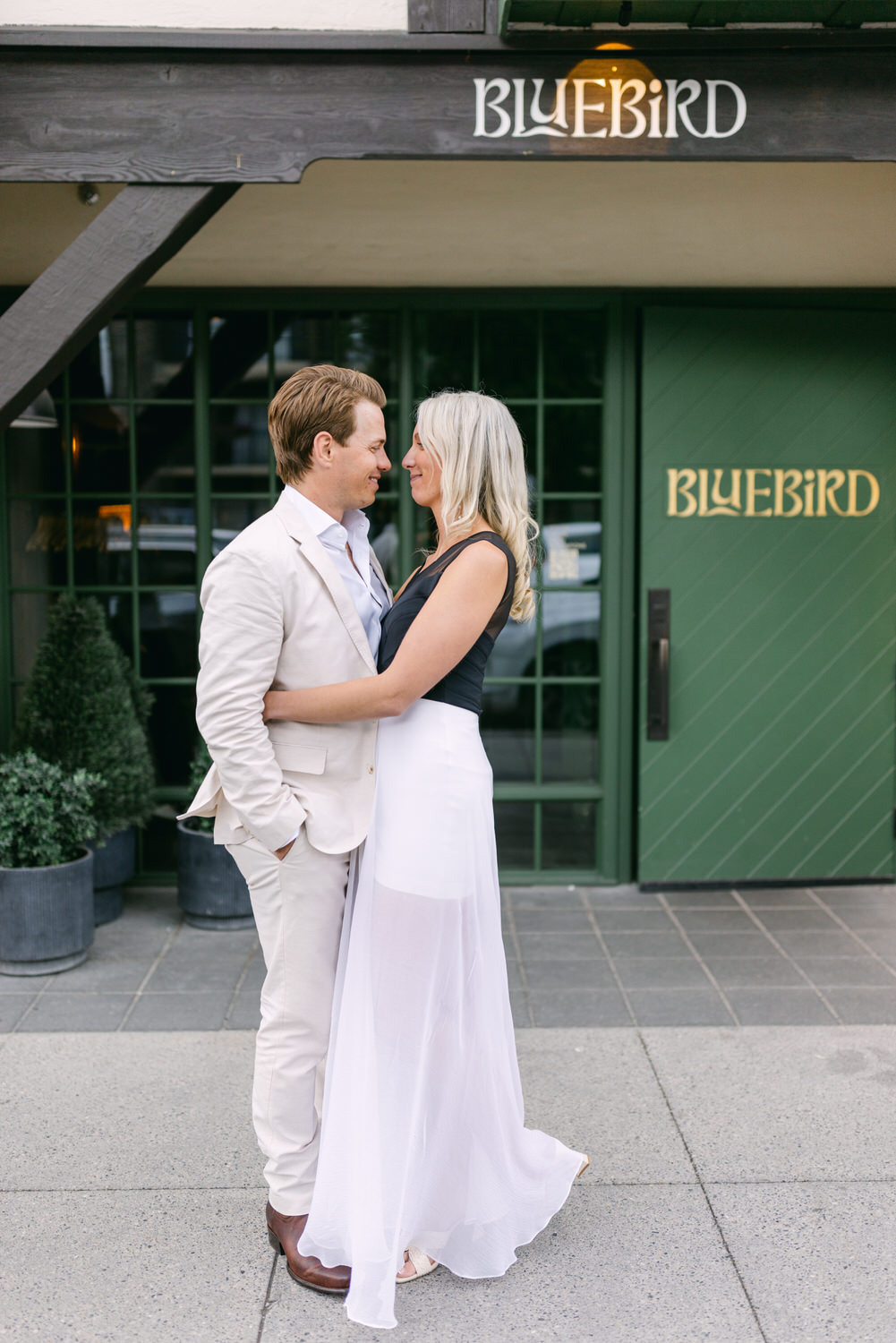 A man in a white suit and a woman in a white dress lovingly embracing in front of the Bluebird Cafe entrance.