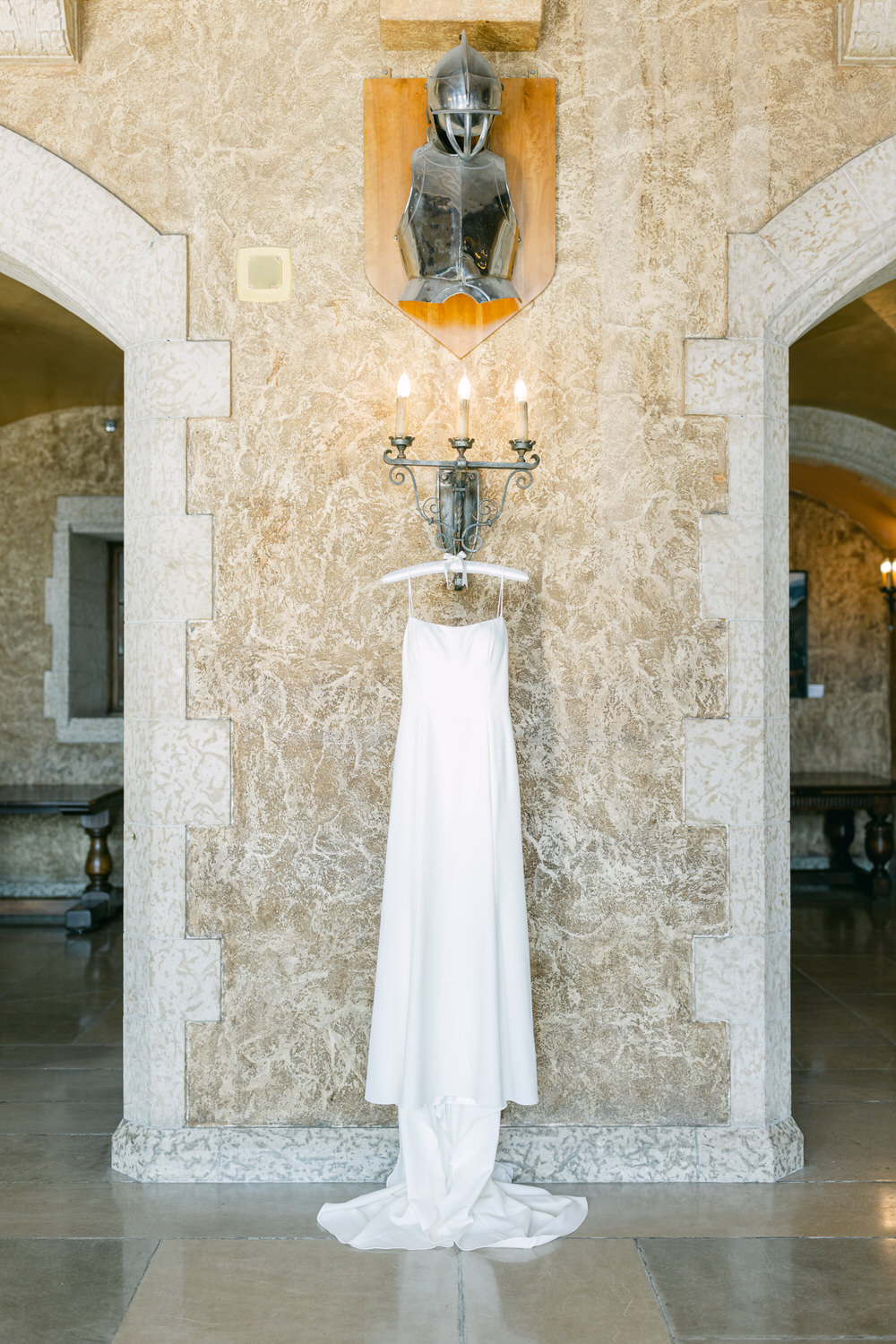 A white bridal dress hanging from a wall sconce beneath an armored knight display on a textured stone wall in a historic building.