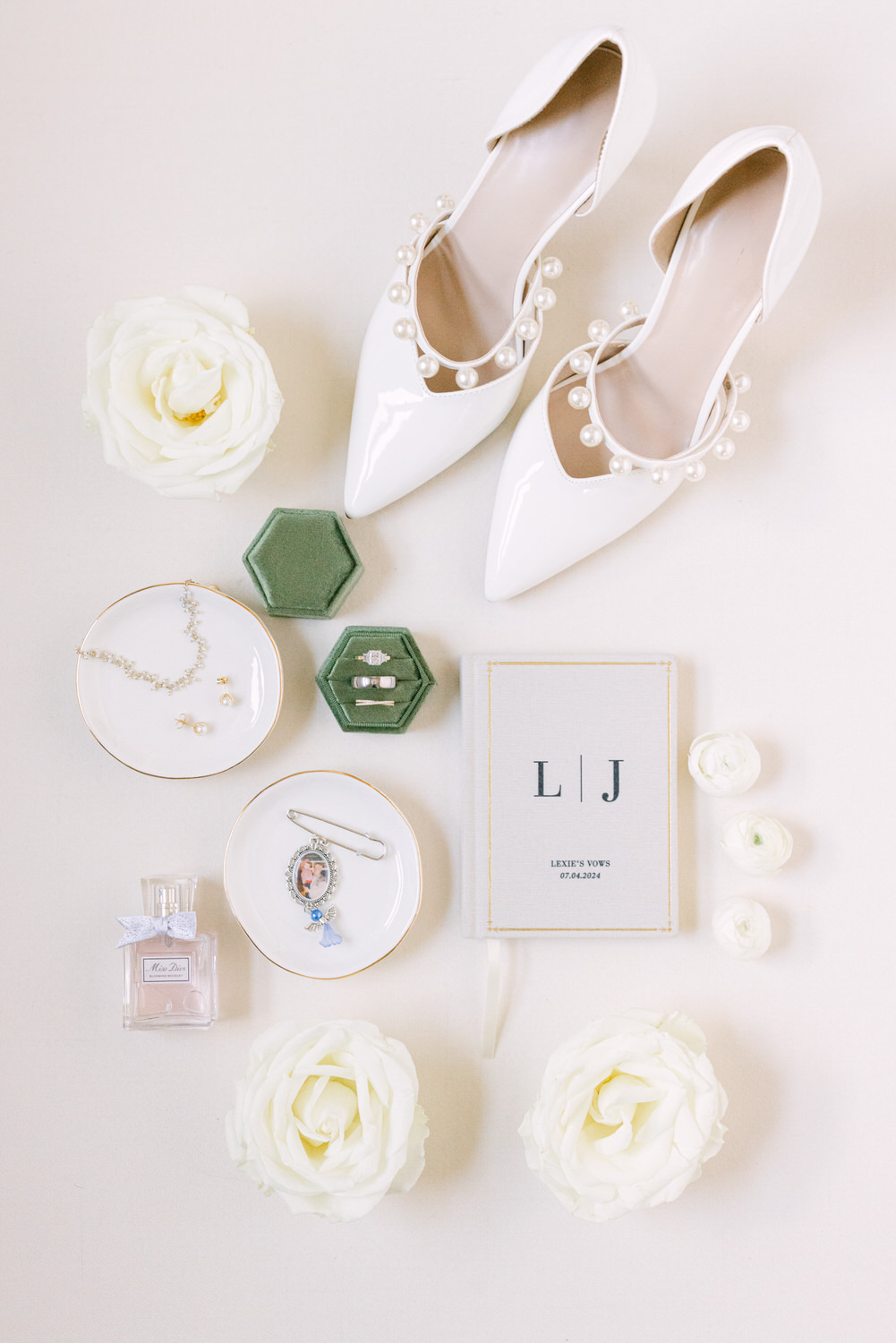 Flat lay of bridal accessories including white pearl-adorned shoes, jewelry, a personalized vow book, and flowers.