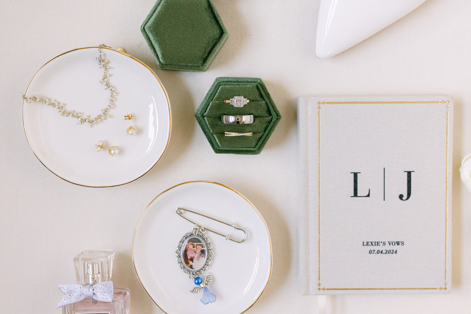 A flat lay of bridal accessories including a necklace, earrings, rings in a green hexagonal box, a brooch with a photo, perfume, and a vow book with the initials L | J on it.