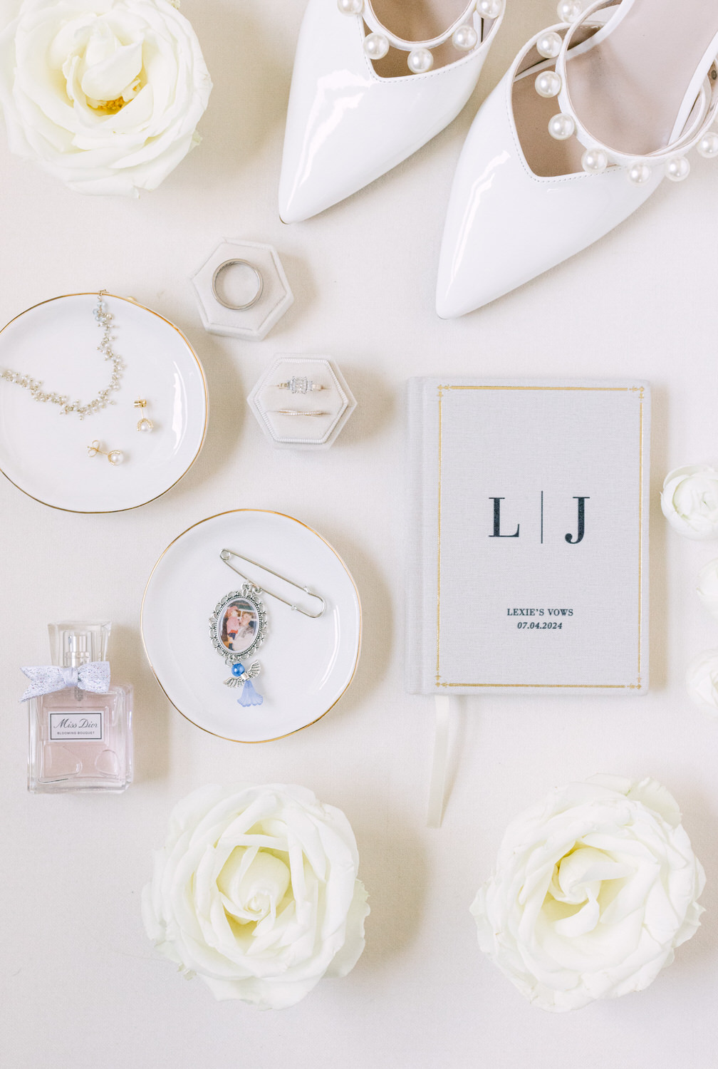 Flat lay of bridal accessories including white shoes, jewelry, perfume, vow book, and roses on a light background