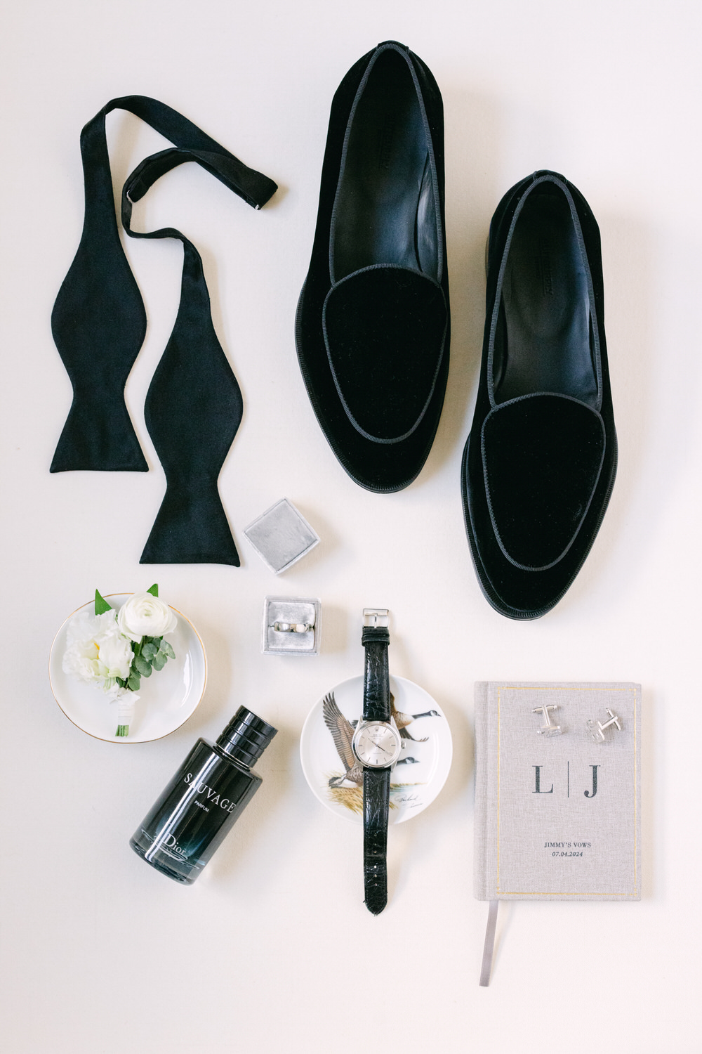 A top view of groom's accessories including black shoes, bow tie, cologne, watch, cufflinks, and a vow book on a light background.