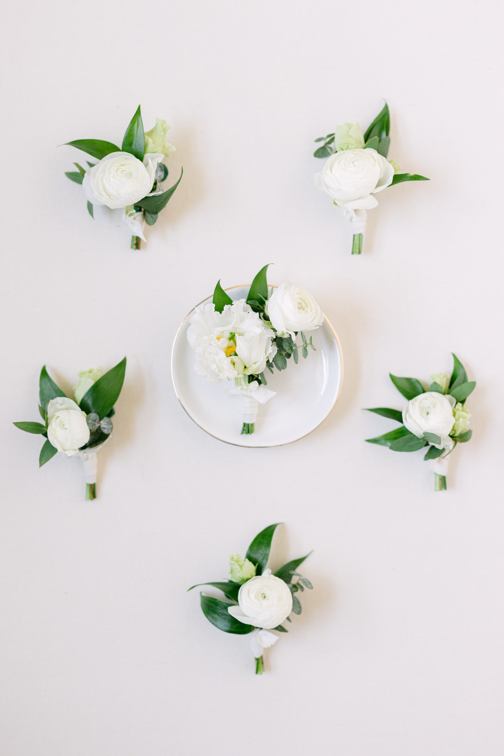 Overhead view of six white wedding boutonnieres and a coordinated floral arrangement placed symmetrically on a white background.