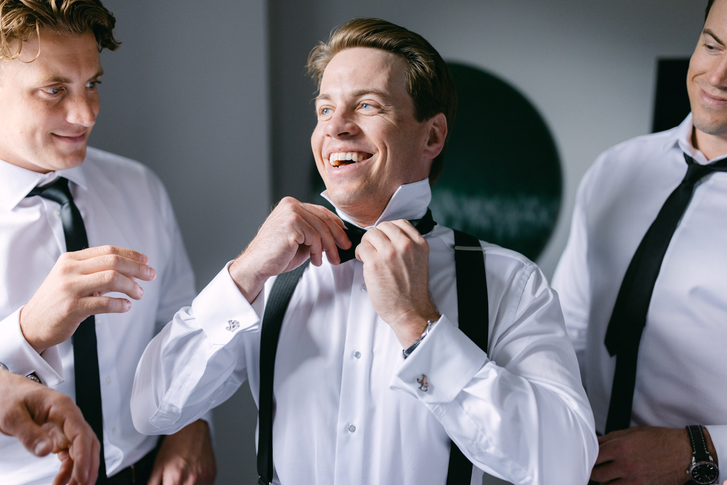 Three men in formal attire with one man adjusting his tie while smiling, surrounded by his friends.