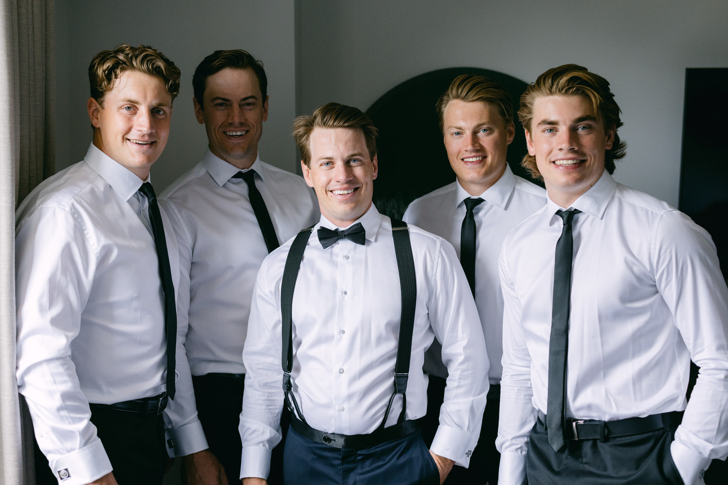 Five smiling men dressed in white shirts, suspenders, and ties, posing confidently.