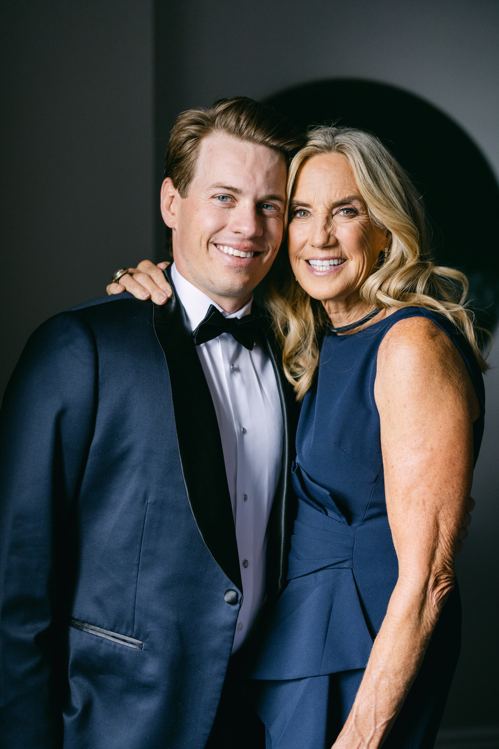 A man in a tuxedo and a woman in a navy blue dress smiling for a portrait with a dark background.