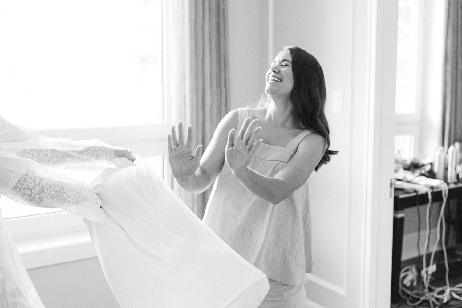 A happy bride in a sundress laughs as she holds up her wedding dress indoors in a monochrome photograph.