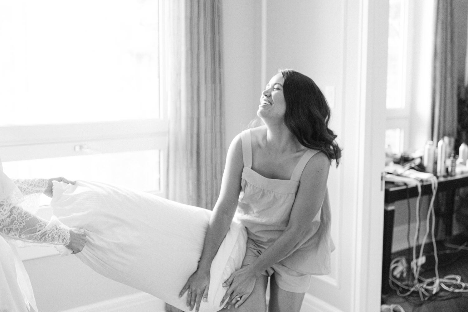 A woman in a sundress laughing joyfully as she holds up a bridal gown in a well-lit room.