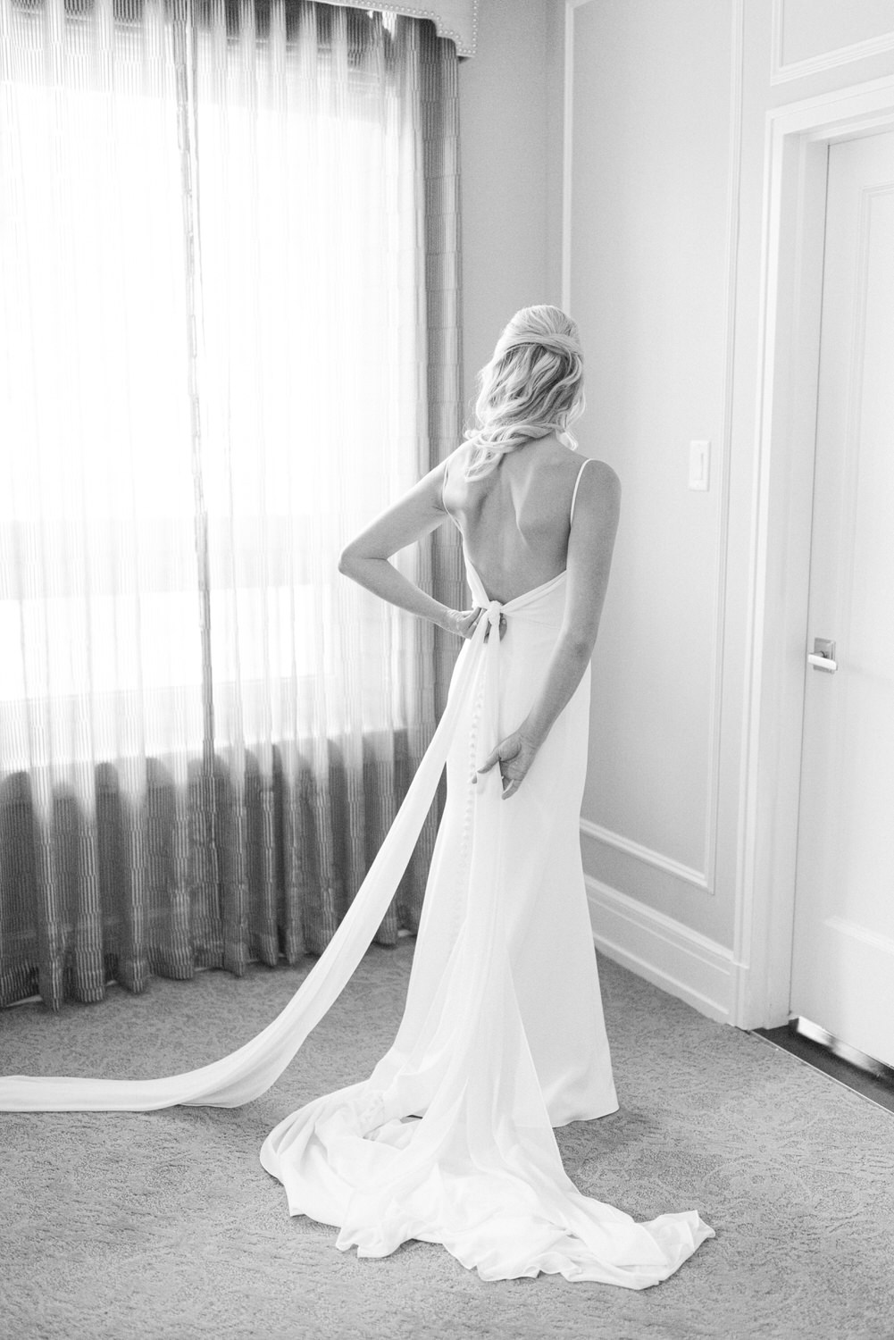 A black and white image of a bride from behind as she adjusts her dress, standing in a room with curtains and carpeted floor.