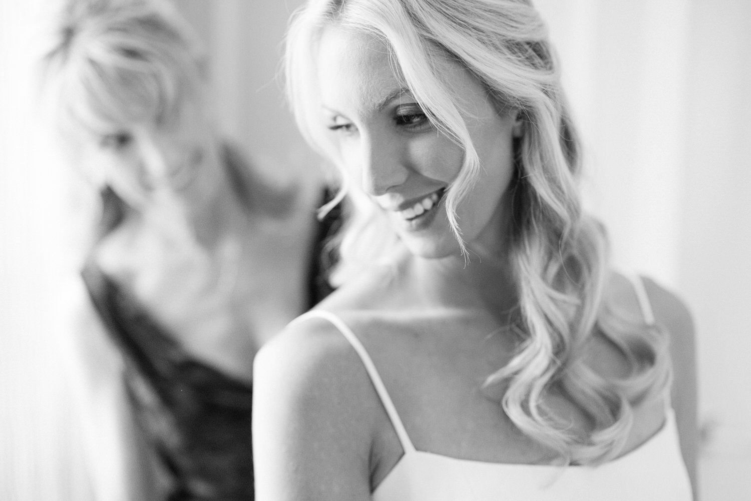 A black and white photo of a smiling woman with flowing hair, standing in focus with another woman reflected in a mirror in the background.