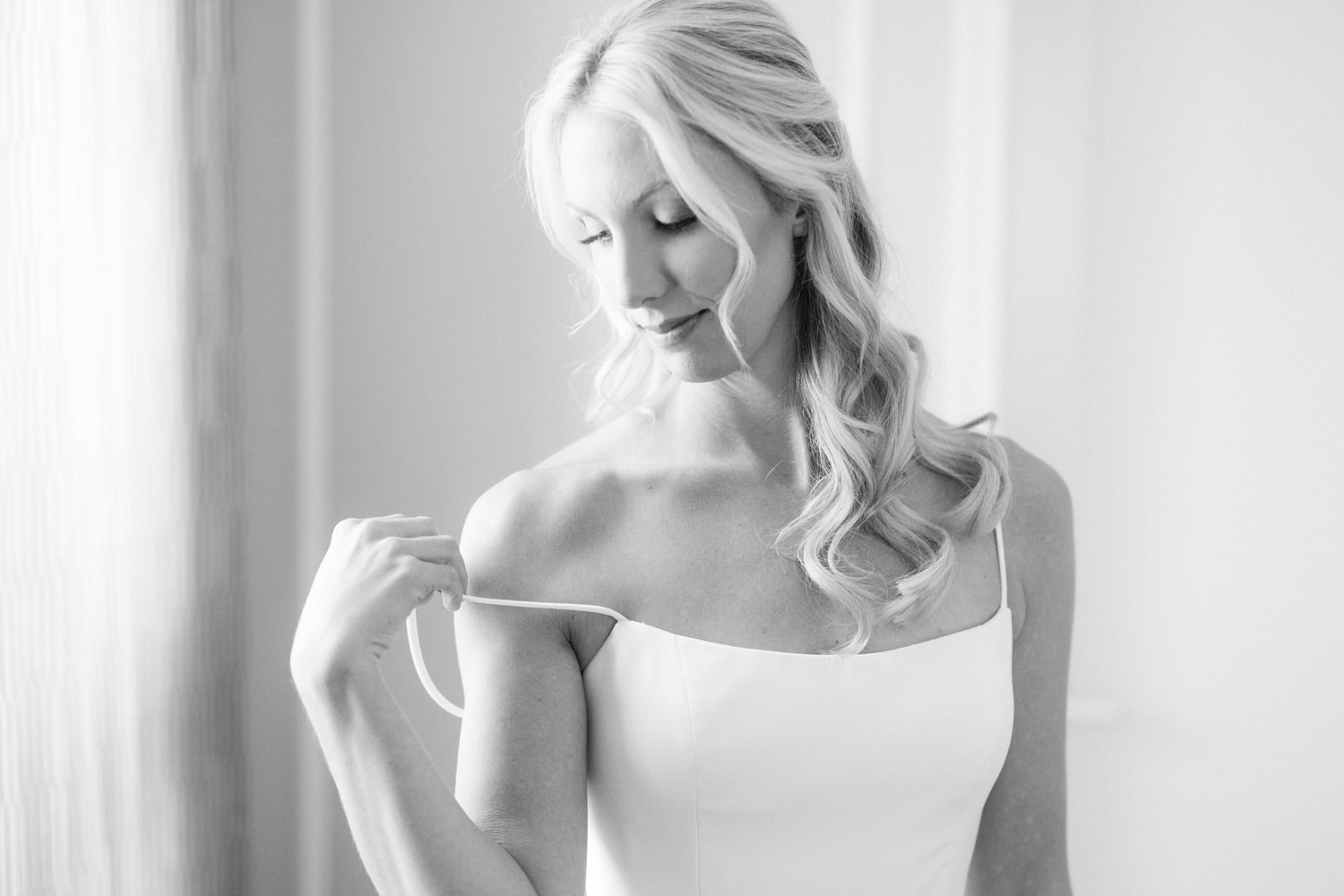 A black and white photo of a woman in a bridal dress, looking down and adjusting her dress, exuding elegance and tranquility.