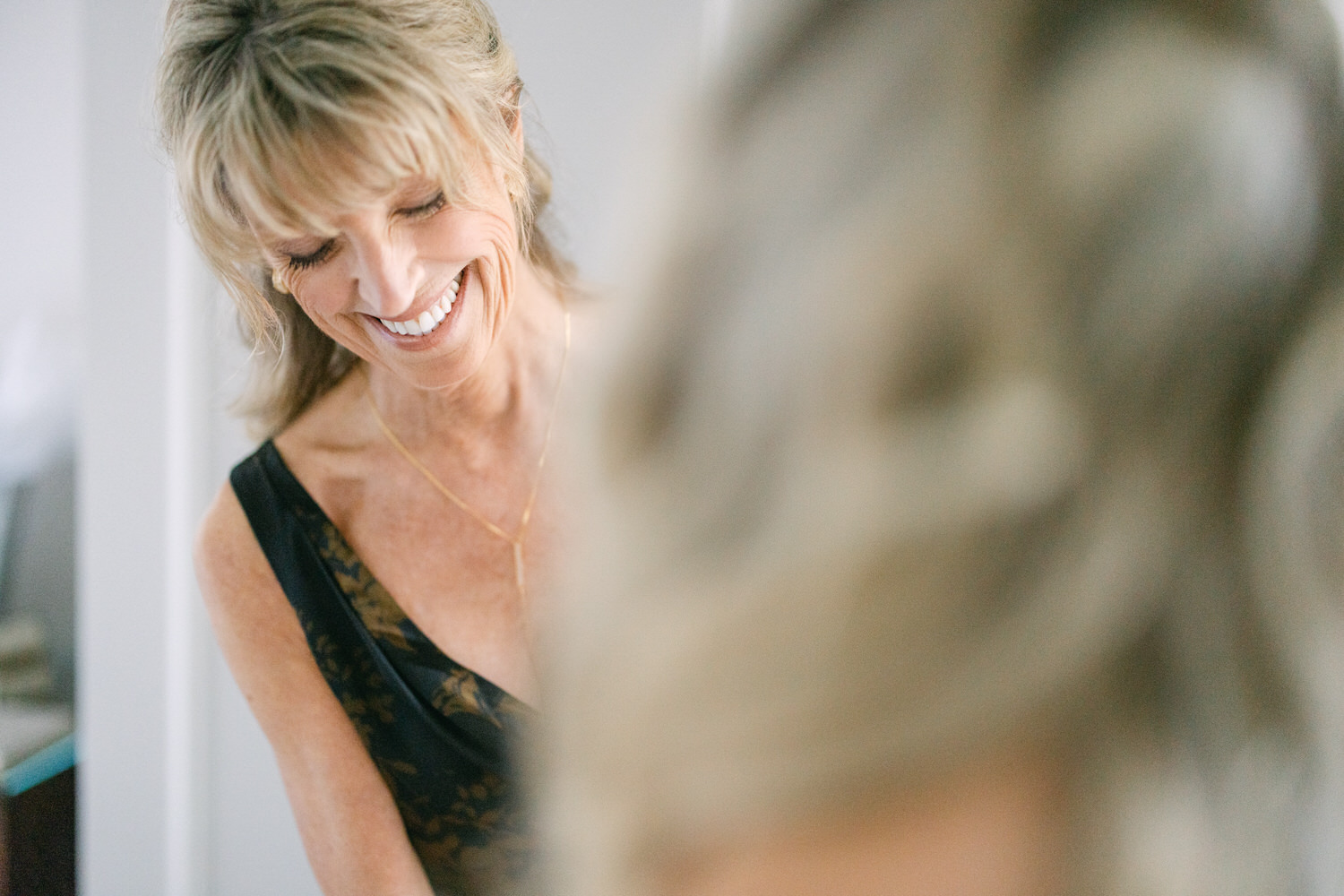 A woman with a radiant smile wearing a black dress with lace details looking down and to the side in a softly lit room.