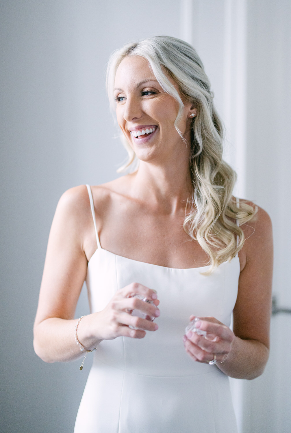 A happy woman with blonde hair wearing a white dress, smiling and looking away, with a light interior background.