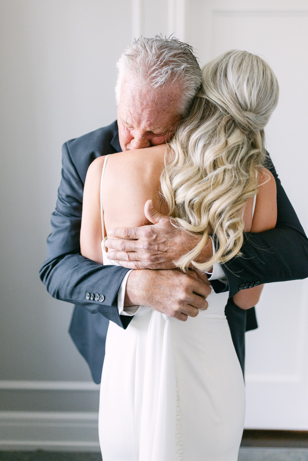 An older man with gray hair in a suit hugs a younger woman with blonde hair in a white dress, expressing a moment of emotional connection.