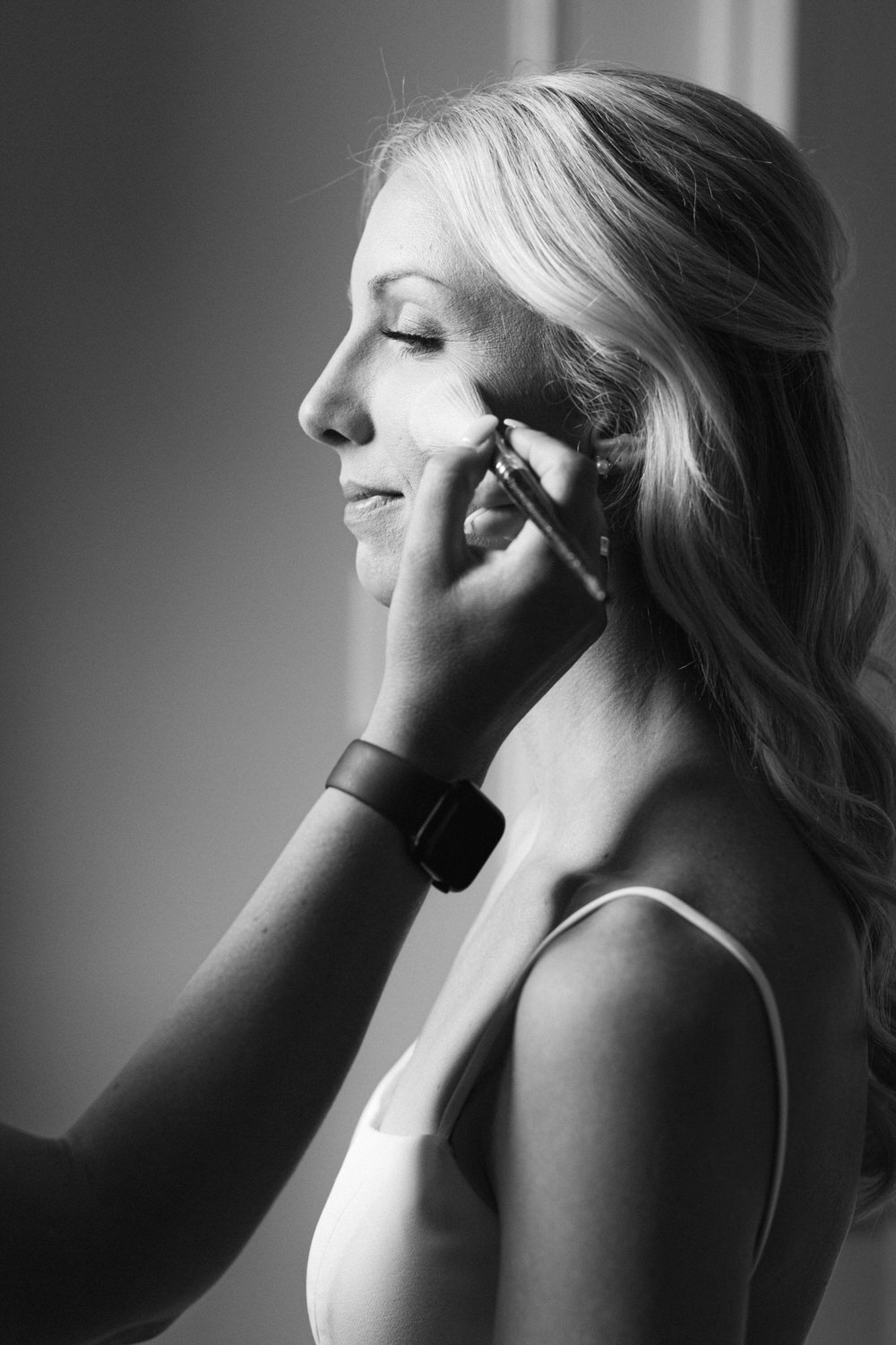 A black and white image of a woman having her makeup applied, eyes closed in a serene expression, wearing a sleeveless top and a smartwatch.