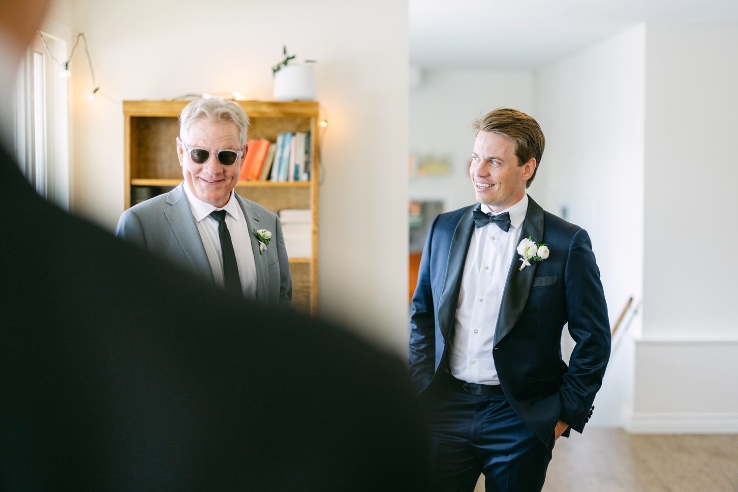 Two men in formal attire smiling at each other in a well-lit room, with the older man wearing sunglasses.