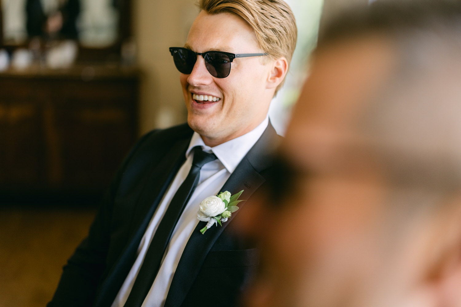 A smiling man in sunglasses and a formal suit with a boutonniere, happily engaged in conversation at an event.