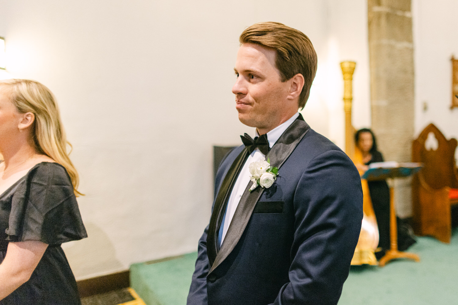 A man in a tuxedo with a boutonniere glancing over his shoulder in a church setting.