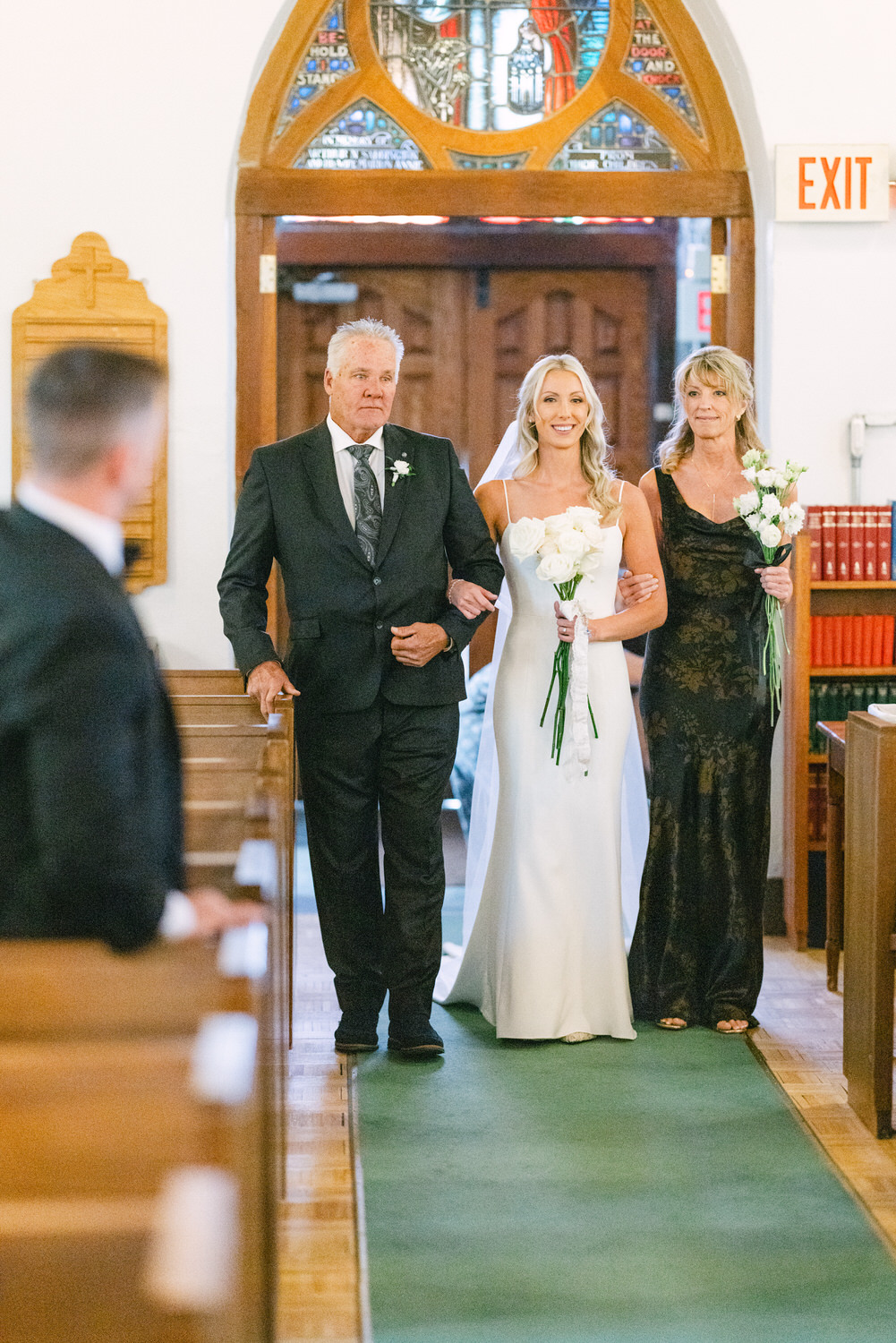 A bride smiling while walking down the aisle flanked by her father and mother in a church setting, with stained glass windows in the background.