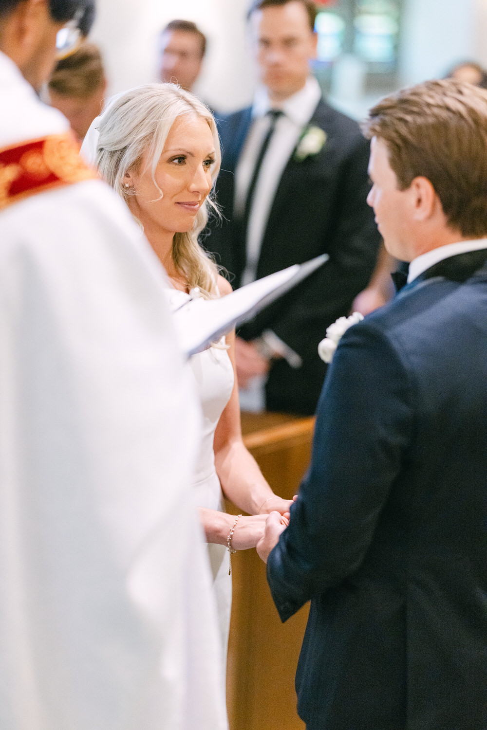 Bride and groom holding hands exchanging vows with officiant in foreground and guest in background.