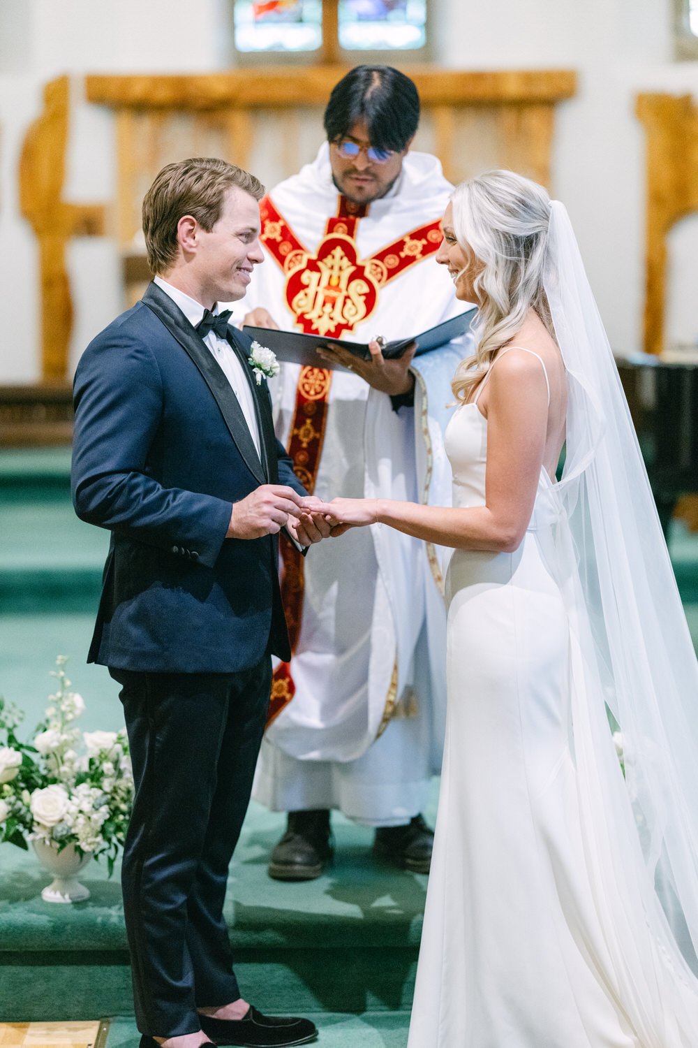 A bride and groom holding hands while exchanging vows in a church ceremony, with a priest standing behind them.