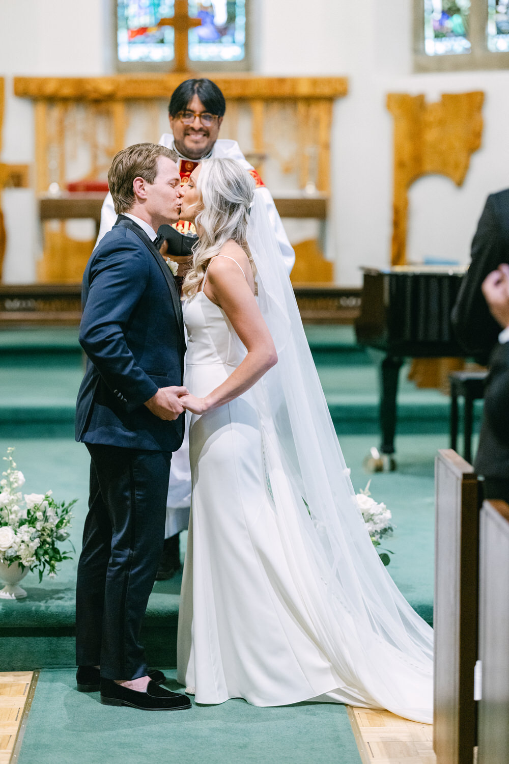 A bride and groom sharing a kiss at the altar with a clergyman smiling in the background, in a church setting with stained glass windows.