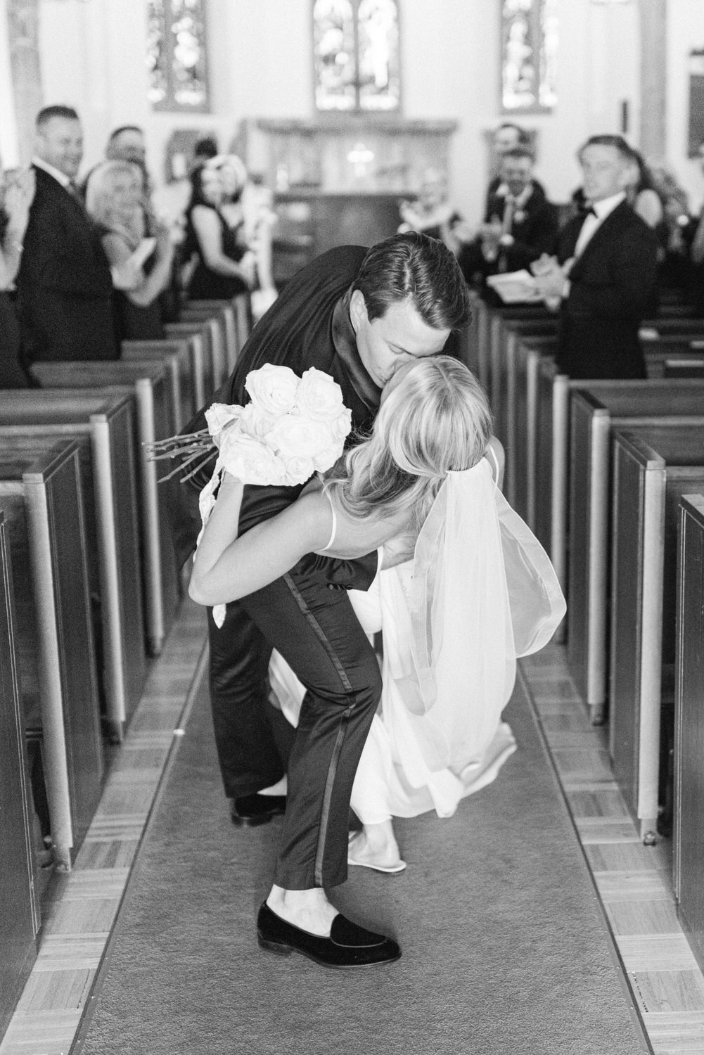 A black and white photo of a groom bending down to kiss a child holding flowers in a church aisle, with guests looking on.