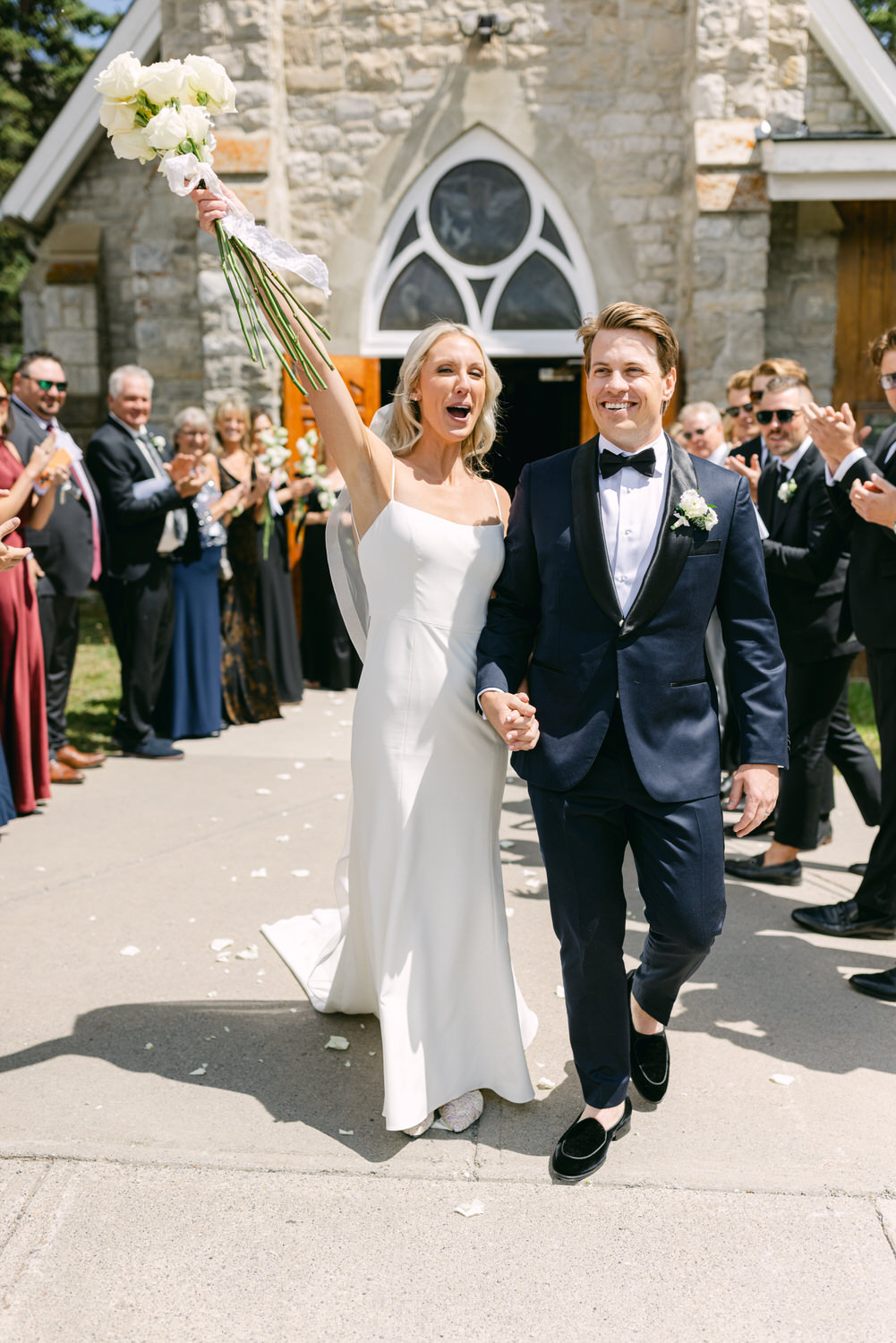 A bride and groom are smiling and holding hands while walking out of a church, with the bride holding up a bouquet, and guests applauding in the background.