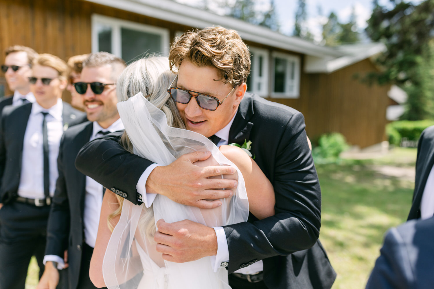 A smiling groom in sunglasses hugging a bride with a veil outdoors, with groomsmen in the background.