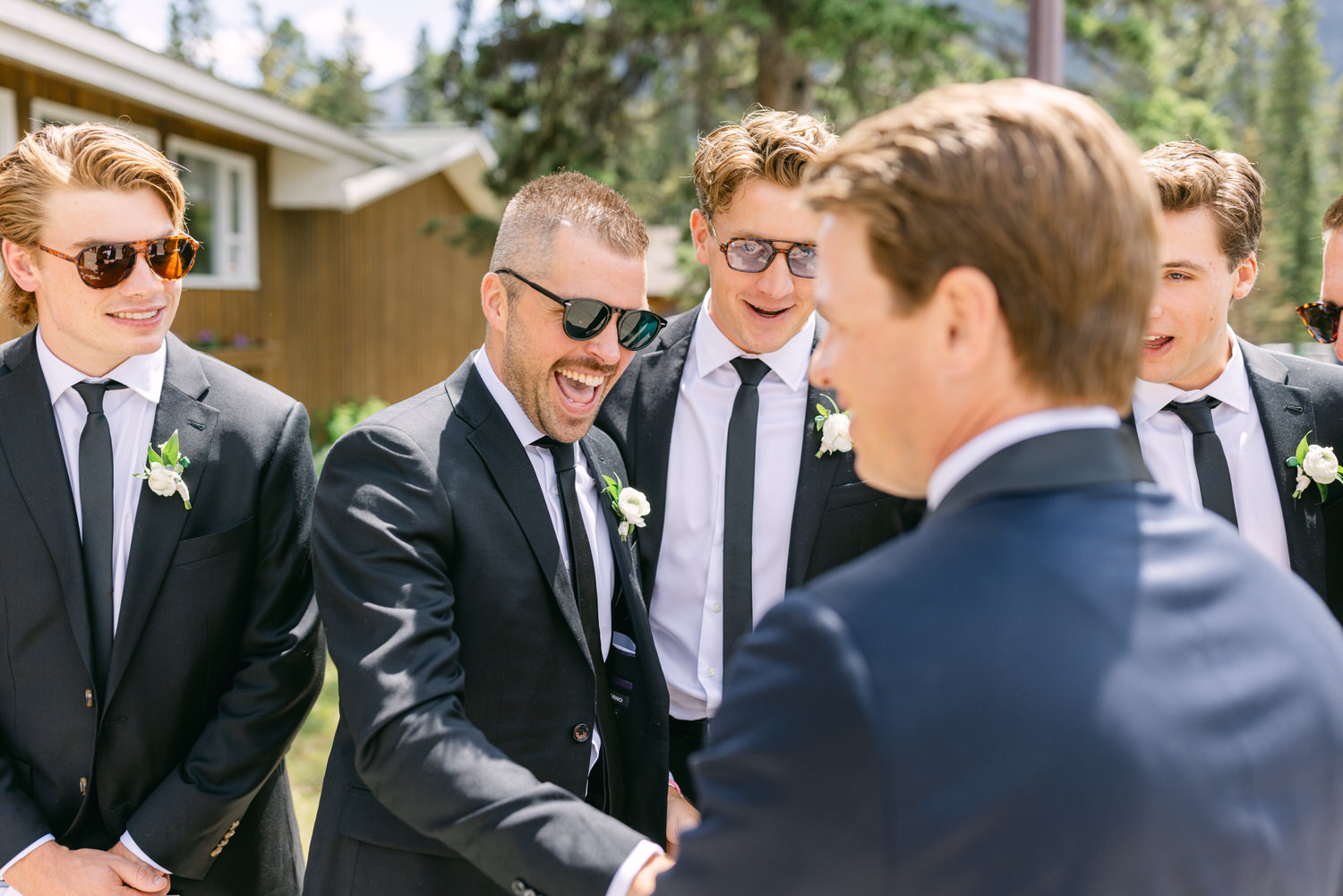 A group of groomsmen laughing and smiling in suits with boutonnieres, sharing a happy occasion outdoors.