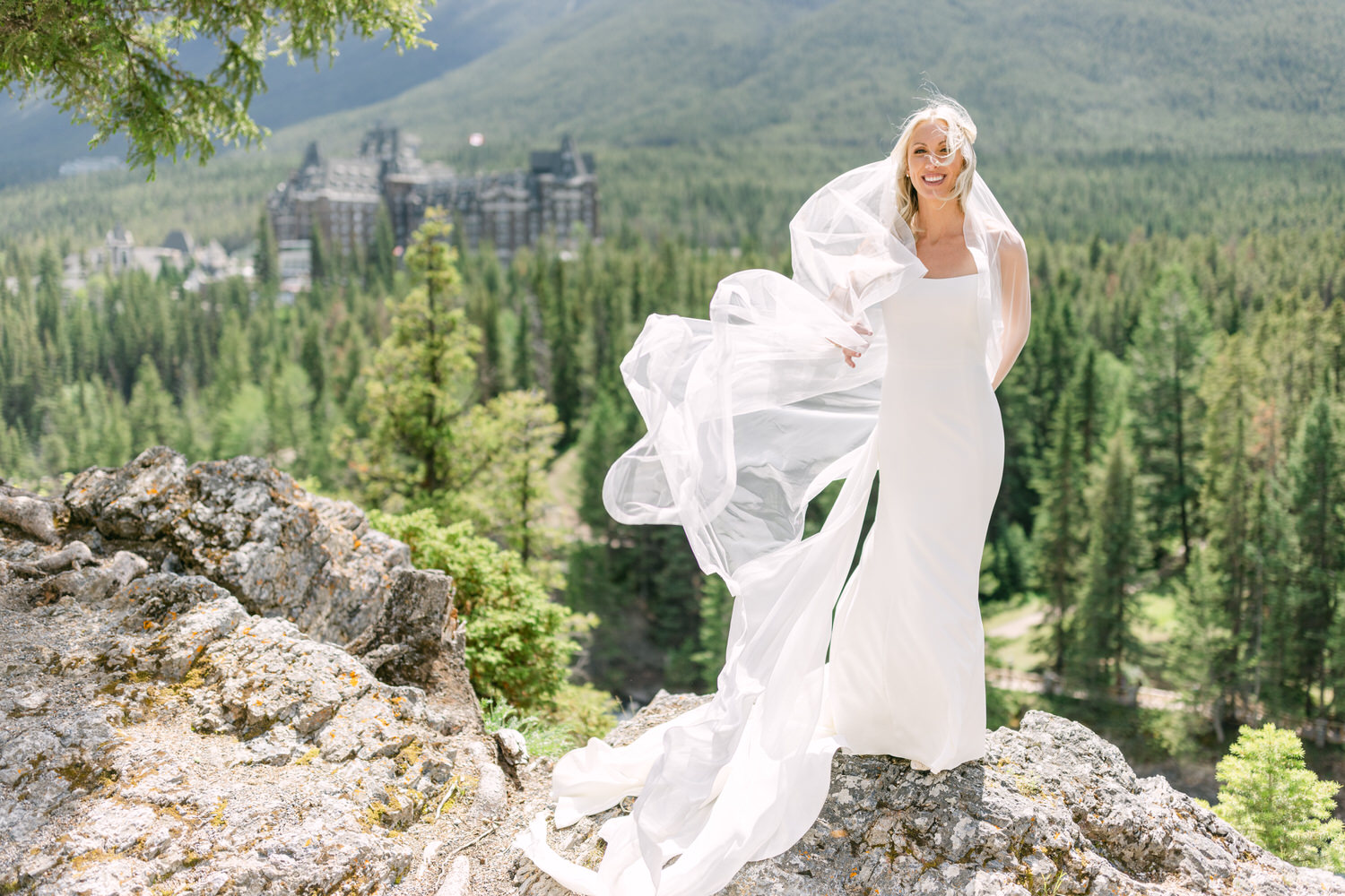 A bride in a white dress with a flowing veil standing on a mountain with a forest and a resort in the background.